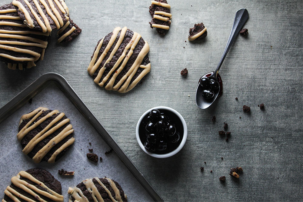 Triple Chocolate Cherry Cookies covered in cherry white chocolate ganache on a cookie sheet with a bowl and spoon of Amarena Cherries