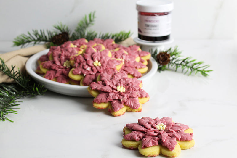 Intricately frosted Poinsettia Sugar Cookies with a jar of Pomegranate Flavor Paste and pine needles in the background