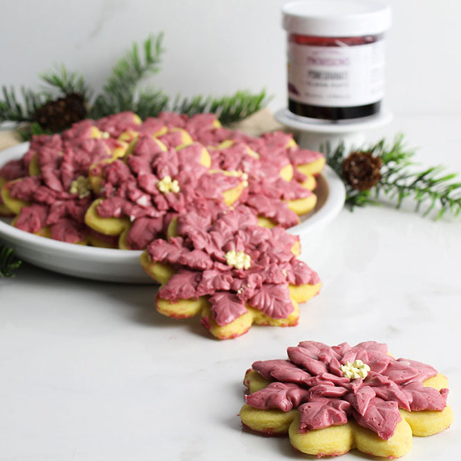 Intricately frosted Poinsettia Sugar Cookies with a jar of Pomegranate Flavor Paste and pine needles in the background