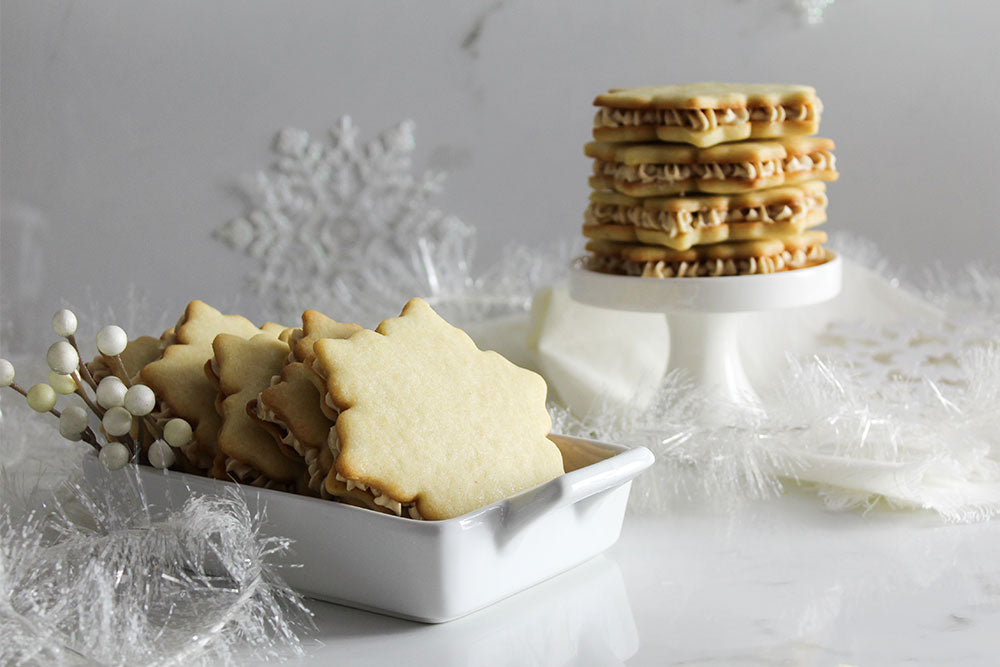 Snowflake vanilla sugar cookies in a loaf pan and stacked on a cake stand in a wintery scene with silver garland, white berries and snowflakes