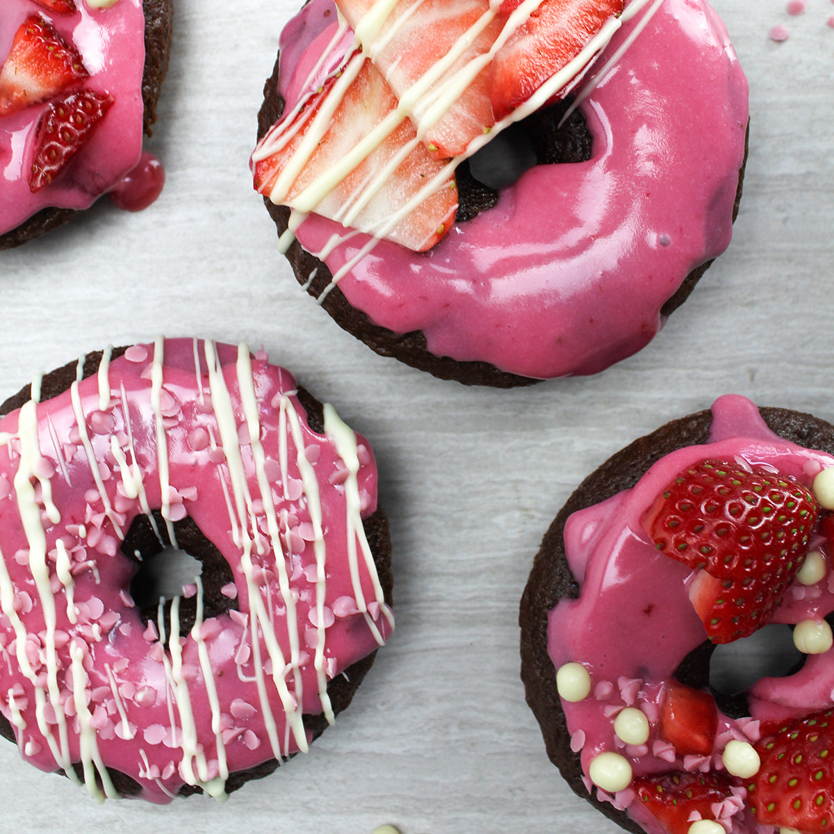 Chocolate cake donut topped with strawberry flavored donut icing, strawberries, pink white chocolate micro drops and white chocolate drizzle